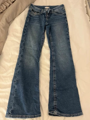 Blå bootcut jeans med broderade fickor - Säljer ett par blå bootcut jeans med snygg slitning och broderade bakfickor. Jeansen har låg midja, klassisk femficksmodell och är tillverkade i denim. Perfekta för dig som gillar en avslappnad men trendig look.