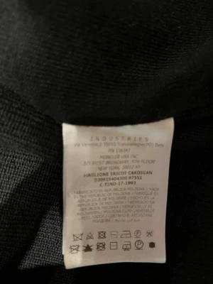Svart cardigan med huva från Moncler - Svart cardigan från Moncler med ribbstickad struktur och huva i kontrasterande material. Långa ärmar och ribbade muddar ger en clean look. Materialet är en mix av ull och polyamid, vilket gör den både mjuk och slitstark. Perfekt för dig som gillar exklusiva detaljer. Storlek 3, passar M-L!