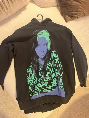 Svart hoodie med grön grafisk print - Cool svart hoodie med stor grafisk print i grönt och blått på framsidan billie Elish.. Hoodien har huva och ribbade muddar vid ärmslut och nederkant. Perfekt för dig som gillar streetwear och vill sticka ut med en unik design. Storlek 158/164 