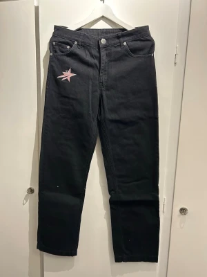 Svarta Lukamachain jeans med rosa tryck - Unika svarta jeans med raka ben och hög midja. På framsidan finns en rosa stjärna och på baksidan står det 'CHAIN WEAR' i stora rosa bokstäver. Jeansen har klassisk femficksdesign och silverfärgade nitar. Perfekta för dig som vill sticka ut.