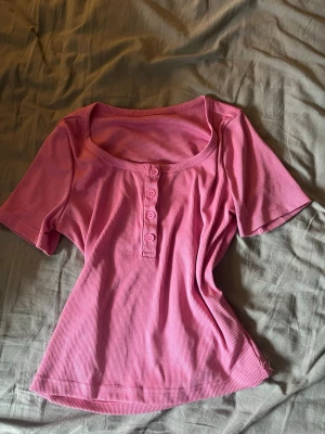 Rosa topp med knappar - Snygg rosa topp med korta ärmar och rundad halsringning. Toppen har dekorativa knappar framtill och är gjord i ett mjukt, stretchigt material som sitter bekvämt. Perfekt att styla med jeans eller kjol för en trendig look.
