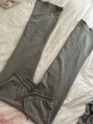 Grå mjukisbyxor med dragsko - Säljer ett par klassiska grå mjukisbyxor med raka ben och dragsko i midjan. Byxorna har en enkel design och är tillverkade i mjukt, stretchigt material som känns skönt mot huden. Finns slitage 