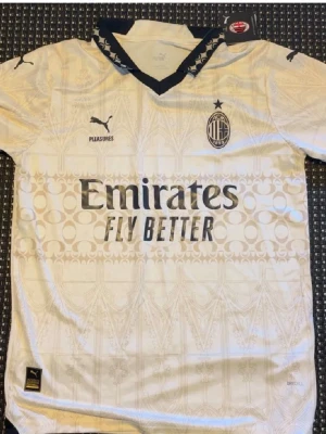 AC Milan bortatröja Puma vit - Snygg vit AC Milan bortatröja från Puma med subtilt mönster, svarta detaljer vid kragen och klubbmärke på bröstet. Tryck från Emirates och wefox. Perfekt för fotbollsträning eller att bära till vardags.