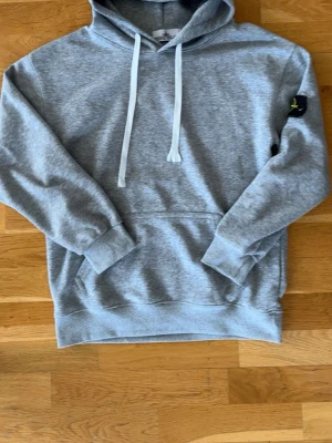 Grå hoodie från Stone Island - Snygg grå hoodie från Stone Island med klassisk huva och vita dragsnören. Tillverkad i mjukt material som känns skönt mot huden. Perfekt för dig som gillar stilrena och avslappnade plagg med en ikonisk logga. Storlek M men passar XS-S