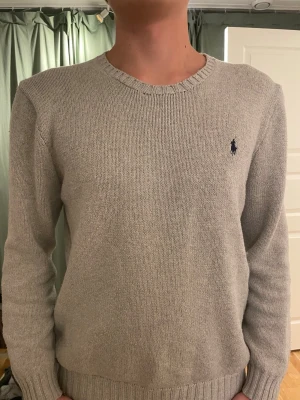 Ljusgrå stickad tröja från Polo Ralph Lauren - Klassisk ljusgrå stickad tröja från Polo Ralph Lauren med rund halsringning och diskret mörkblå logga broderad på bröstet. Tröjan har ribbade muddar vid ärmslut och nederkant, perfekt för en clean och stilren look.