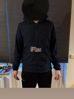 Marinblå vindjacka Tommy Hilfiger - Snygg marinblå vindjacka från Tommy Hilfiger Denim med huva och halv dragkedja framtill. Jackan har en stor ficka med logga på bröstet och en liten flagga på ärmen. Perfekt för blåsiga dagar och enkel att matcha med olika outfits.