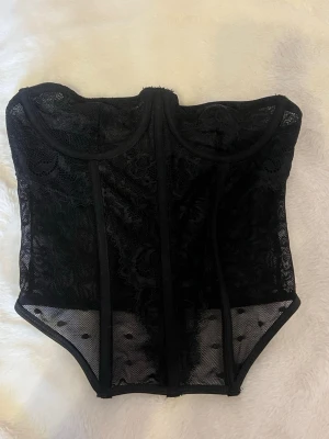 Svart spetskorsett  - Snygg svart korsett med genomskinliga meshpartier och spetsdetaljer. Har markerade sömmar och bygel för extra stöd samt hakknäppning bak