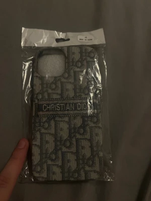 iPhone 13 Christian Dior grå mobilskal  - Snyggt mobilskal från Christian Dior med klassiskt mönster i ljusgrått och mörkare grå. Skalet har en textil känsla och logotypen broderad på baksidan. Passar dig som vill ha en exklusiv look till mobilen.