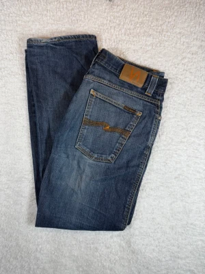 Nudie Jeans Slim Jim W36 L 32 - Säljer ett par klassiska blå jeans från Nudie Jeans med raka ben och snygga slitningar. Jeansen har fem fickor, kontrastsömmar och en brun läderpatch med logga bak i midjan. Tillverkade i bomull med en cool tvättad look.
