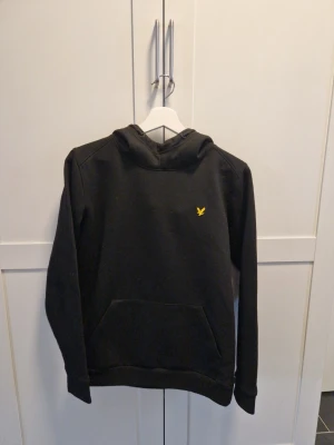 Svart hoodie från Lyle & Scott - Snygg svart hoodie från Lyle & Scott med klassisk gul logga på bröstet. Hoodien har huva och en stor känguruficka framtill. Tillverkad i mjukt material som känns skönt mot huden. Perfekt för en avslappnad och stilren look.