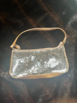 Guldig paljettväska från Vezzano - Säljer en snygg handväska från Vezzano med guldiga paljetter på framsidan och matchande guldfärgat tyg. Väskan har en enkel axelrem och dragkedja upptill. Perfekt för dig som vill sticka ut med lite extra glitter. Lite fläckar på insidan som syns på bild 2 