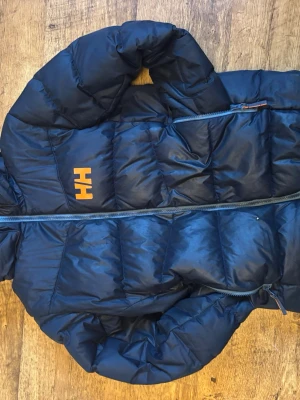 Mörkblå Helly Hansen dunjacka - Jag säljer en riktigt snygg mörkblå dunjacka från Helly Hansen med huva och orange logga på bröstet och ryggen. Jackan har quiltad design och dragkedja framtill. Perfekt för kyliga dagar och enkel att matcha med olika outfits. En riktigt varm jacka dock väldigt välanvänd med flera slitage perfekt jacka att leka i som man inte behöver vara rädd om.