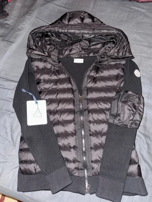 Svart hoodie-cardigan med dragkedja - Snygg svart cardiganjacka med vadderad front och huva. Jackan har dragkedja, långa ärmar och en ficka på ena ärmen. Perfekt för lager-på-lager och streetwear-stil. Materialet är en mix av  tyg och mjuk bomull.