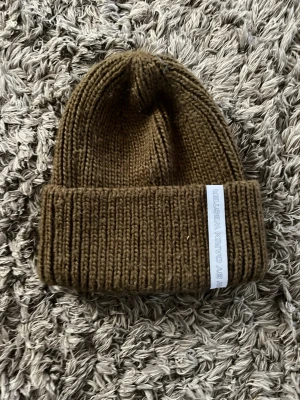 Grön stickad mössa - Säljer en grön, ribbstickad mössa från Carin Wester. Mössan har uppvikt kant och en vit etikett med logga på sidan. Perfekt för dig som vill ha en enkel men snygg accessoar till hösten, våren och vintern.