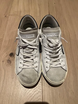 Vita Philippe Model sneakers - Snygga vita sneakers från Philippe Model med grå mockadetaljer på tån och svart hälparti. Klassisk låg modell med vita skosnören och diskret logga på sidan. Skorna är tillverkade i läder och mocka för en stilren och trendig look.