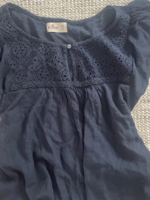 Mörkblå blus från Hollister - Mörkblå blus från Hollister i storlek XS. Blusen har korta ärmar, rund halsringning och ett dekorativt broderat parti med hålmönster upptill samt en liten knapp framtill. Tillverkad i ett lätt, luftigt tyg som ger en avslappnad look.