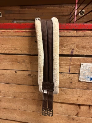 Brun sadelgjord med fluff - Brun sadelgjord med mjuk, beige fluff på och metallspännen i ändarna. Perfekt för att ge extra komfort till hästen och minska skav. Tillverkad i slitstarkt tyg och syntetmaterial. Str: 120cm. Använt men i gott skick!