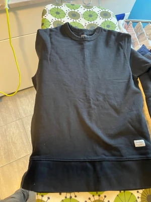 Svart sweatshirt från Les Deux - En stilren svart sweatshirt från Les Deux med rund halsringning och ribbade muddar vid ärmslut och nederkant. Tröjan har en liten vit patch med logga nere vid fållen. Perfekt för en clean och avslappnad look.
