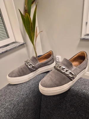 Deabused gråa slip-on sneakers med silver kedjedetalj - Snygga gråa slip-on sneakers i mocka med chunky silverfärgad kedja över vristen och vit platåsula. Insidan är fodrad i brunt läder för extra komfort. Perfekta för dig som vill ha en stilren men ändå unik look.