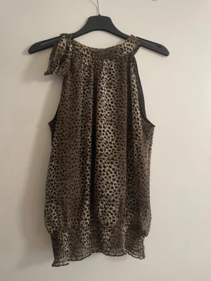 Leopard topp - Säljer en snygg ärmlös topp med leopardmönster och knyt vid axeln. Toppen har en lös passform och resår i nederkanten för en skön siluett. Perfekt för dig som vill sticka ut lite extra. 