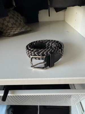 Flätat svart och grått bälte - Snyggt flätat bälte i svart och grått med metallspänne och detaljer i skinn. Perfekt accessoar för att lyfta din outfit och funkar till både jeans och kostymbyxor. Bältet har en flexibel design som gör det enkelt att justera längden.