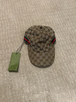 Gucci keps med GG-mönster - Snygg keps från Gucci med klassiskt GG-mönster i beige och brunt. Kepsen har gröna och röda detaljer på sidorna och är tillverkad i canvas med insida i vitt tyg. Perfekt för dig som vill ha en exklusiv och trendig look.