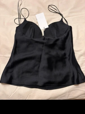 Svart satin linnetopp med tunna band - Snygg svart linnetopp från Zara i glansigt satinmaterial. Toppen har tunna axelband och lätt vadderade kupor för en smickrande passform. Perfekt att styla med jeans eller kjol för en trendig look. Ny med prislapp!