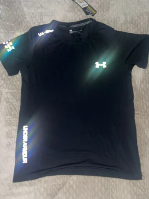 Svart tränings-tshirt från Under Armour - Svart kortärmad tränings-tshirt från Under Armour med vitt logotryck på bröstet och axeln. Materialet är tekniskt och andas, perfekt för löpning eller gym. Rund halsringning och smidig passform. Snygga detaljer med tryck på ärm och nederkant.