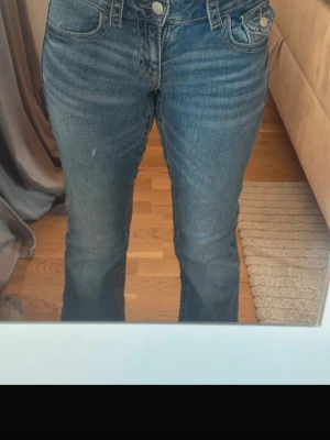 Blå bootcut jeans med dubbla knappar - Säljer ett par blå bootcut jeans med dubbla knappar i midjan och klassiska fickor fram och bak. Jeansen har en lätt tvättad look och sitter snyggt över höfterna. Perfekta för dig som gillar en avslappnad men ändå trendig stil.