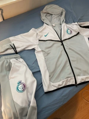 Nike grått träningsset med luva - Säljer ett grått träningsset från Nike med matchande byxor och hoodie. Setet har klubbmärke på både jacka och byxa, svarta detaljer och dragkedja framtill. Materialet är mjukt och stretchigt, perfekt för träning eller uppvärmning.