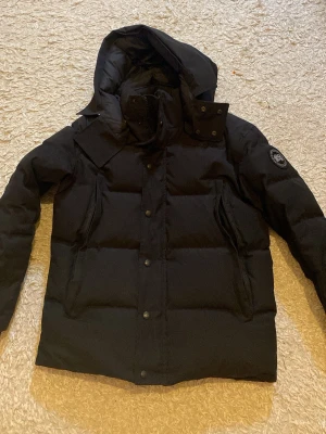 Svart dunjacka från Canada Goose - (Äkta)Säljer en svart dunjacka från Canada Goose med huva och hög krage.har använt jackan i 2 månader. Jackan har dragkedja och knappar framtill, två snedställda fickor och en patch med logga på ärmen. Perfekt för kalla vinterdagar och riktigt varm tack vare dunfyllningen. 