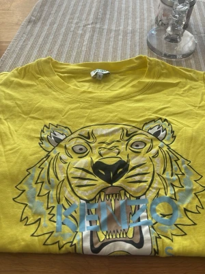Gul Kenzo t-shirt med tigertryck - Säljer en gul t-shirt från Kenzo med det ikoniska tigertrycket framtill och Kenzo-loggan i blått. T-shirten har rund hals och korta ärmar. Perfekt statement-plagg för dig som vill sticka ut med färg och cool design.