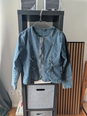 Blå jeansjacka med snörningsdetaljer - Trendig blå jeansjacka med dragkedja framtill och unika snörningsdetaljer på både fram- och baksidan. Jackan har rund halsringning och långa ärmar. Perfekt för dig som vill ha en lite annorlunda jeansjacka med coola detaljer.