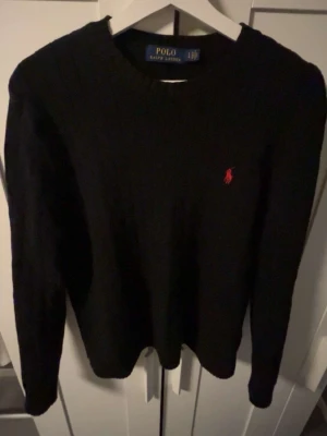 Svart stickad tröja från Polo Ralph Lauren - Klassisk svart stickad tröja från Polo Ralph Lauren med rund halsringning och den ikoniska röda logotypen broderad på bröstet. Tröjan har lång ärm och ribbade muddar. Perfekt för en stilren och enkel look.