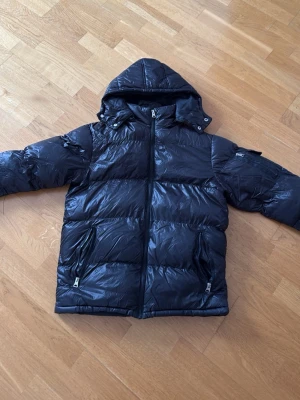Svart pufferjacka med huva - Snygg svart pufferjacka med glansig finish och avtagbar huva. Jackan har två dragkedjeförsedda fickor framtill och quiltad design. Perfekt för kalla vinterdagar och ger en modern streetwear-look.