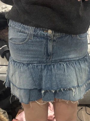 Blå jeanskjol med volanger - Säljer en kort jeanskjol i blå denim med två lager volanger och råa kanter för en edgy look. Kjolen har klassiska fickor fram och bak samt knapp och dragkedja i midjan. Perfekt för dig som gillar en avslappnad men trendig stil. kids 170 ~ xs/s