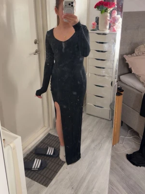 Svart långklänning med slits - Snygg svart långklänning med lång ärm och djup slits på sidan. Klänningen har en tight passform och är perfekt för dig som vill ha en stilren och elegant look. Materialet känns mjukt och följsamt mot kroppen.
