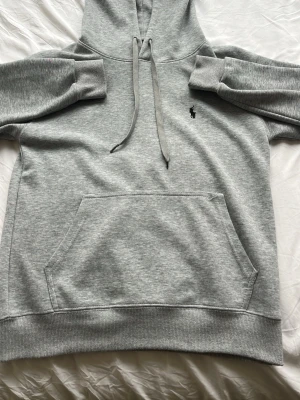 Grå hoodie från Polo Ralph Lauren - Klassisk grå hoodie från Polo Ralph Lauren med svart broderad logga på bröstet. Hoodien har stor känguruficka, ribbade muddar och dragsko i huvan. Tillverkad i mjukt bomullsmaterial som är perfekt för chill dagar. Lite trasig i fickan som inte syns alls när man har på sig den.