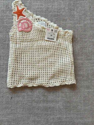 Virkat linne med applikationer från Zara - Ett krämvitt virkat linne från Zara med asymmetrisk axel och dekorativa applikationer i form av en orange stjärna och en rosa spiral. Linnet har vågig kant och är gjort i ett luftigt, nätliknande material. Perfekt för en unik och trendig stil.