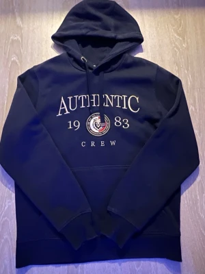 Mörkblå hoodie AUTHENTIC 1983 - Snygg mörkblå hoodie med huva och dragsko. På bröstet står det 'AUTHENTIC 1983 CREW' med ett broderat emblem i vitt, rött och blått. Hoodien har en klassisk känguruficka och ribbade muddar vid ärmar och nederkant. Perfekt för en chill och sportig look.