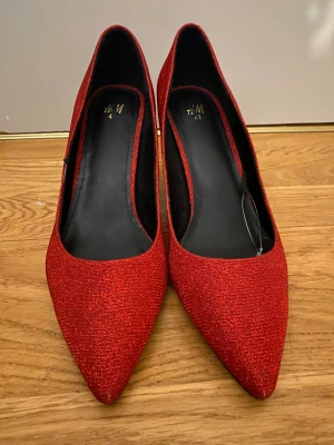 Röda glittriga pumps från H&M - Snygga röda pumps från H&M med glittrigt tyg och spetsig tå. Skorna har en klassisk klack och svart innersula, perfekta för att sticka ut. Materialet är textil med skimrande finish.