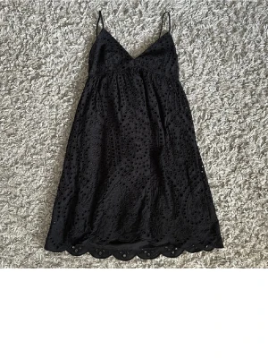 Svart broderad kort klänning - Svart kort klänning med tunna axelband och v-ringning. Klänningen har broderade detaljer och en vågig kant nertill. Baksidan har snörning för extra detalj. Perfekt för dig som gillar en feminin och stilren look.