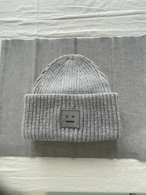Grå ribbstickad mössa Acne Studios - Säljer en grå ribbstickad mössa från Acne Studios med det ikoniska ansiktslogotypen framtill. Mössan är i tjockt, mjukt material och har bred uppvikt kant. Perfekt för kalla dagar och ger en clean, stilren look.