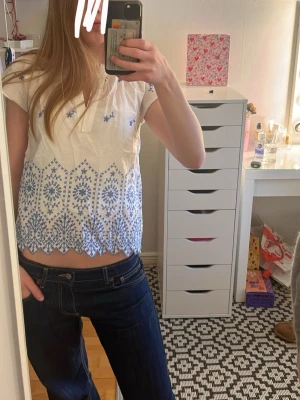 Vit blus med blå broderier - Söt vit blus med blå broderade detaljer och v-ringning. Blusen har korta ärmar och ett fint spetsmönster längst ner. Perfekt till jeans eller kjol för en fräsch look. Materialet känns lätt och luftigt, perfekt för varmare dagar.