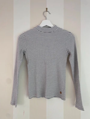 Randig ribbad långärmad polotopp - Snygg vit och svart ribbad polotopp med tunna horisontella ränder. Toppen har lång ärm och en tight passform som sitter snyggt på kroppen. Tillverkad i mjuk bomullsmix som känns skön mot huden. Perfekt att styla med jeans eller kjol. Är i storlek 158/164.