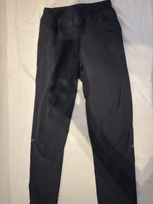 Svarta träningsbyxor med resårmidja - Säljer ett par svarta träningsbyxor med resår i midjan och raka ben. Byxorna har reflexdetaljer runt benen och är tillverkade i ett lätt, funktionsmaterial som passar perfekt för löpning eller gym. Snygg och enkel design utan synligt märke.