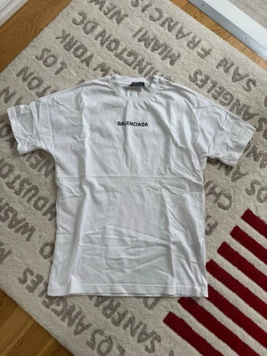 Vit Balenciaga t-shirt med tryck - Vit t-shirt från Balenciaga med svart logotyptryck fram på bröstet och stort tryck på ryggen. Klassisk passform med rund hals och korta ärmar. Tillverkad i mjuk bomull för en skön känsla och enkel stil.
