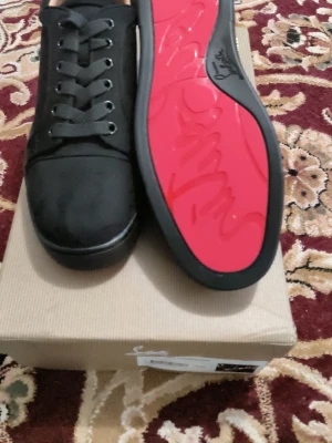 Svarta sneakers från Christian Louboutin - Snygga svarta sneakers från Christian Louboutin med klassisk röd sula och diskret logga. Skorna har en låg profil, svarta skosnören och ovandel i textil eller mocka. Perfekta för dig som vill ha en clean och exklusiv look.