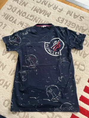 Mörkblå Moncler t-shirt med tryck - Säljer en mörkblå t-shirt från Moncler med stort vitt och rött logotryck på bröstet och mindre tryck över hela tröjan. T-shirten har rund hals och korta ärmar. Perfekt för dig som gillar streetwear och vill sticka ut lite extra.