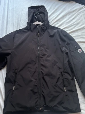 Svart Moncler vindjacka med huva - Svart vindjacka från Moncler med huva och dragkedja framtill. Jackan har två framfickor med dragkedja och en ficka med knapp på ärmen där Moncler-loggan sitter. Tillverkad i ett slätt, vattenavvisande material. Perfekt för blåsiga dagar.